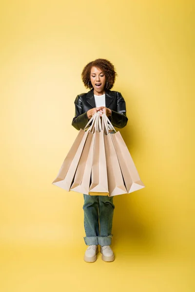 Mulher americana africana animado em jaqueta de couro elegante segurando sacos de compras com compras em amarelo — Fotografia de Stock