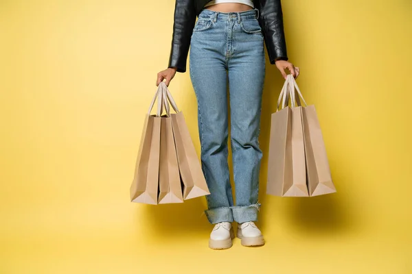 Vista ritagliata di donna afroamericana in giacca di pelle e jeans che tiene borse della spesa con acquisti su giallo — Foto stock