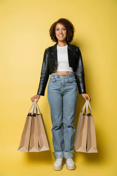 Volle Länge lächelnde afrikanisch-amerikanische Frau in stylischer Lederjacke mit Einkaufstaschen mit Einkäufen auf gelb — Stockfoto