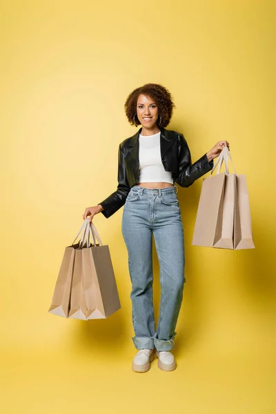 Pleine longueur de femme afro-américaine positive dans une veste en cuir élégant tenant des sacs à provisions avec des achats sur jaune — Photo de stock