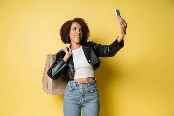 Heureuse femme afro-américaine dans une veste en cuir élégant tenant des sacs à provisions et de prendre selfie sur jaune — Photo de stock