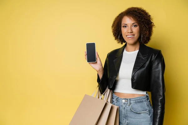Mujer afroamericana feliz en chaqueta de cuero sosteniendo bolsas de compras y teléfono móvil con pantalla en blanco en amarillo - foto de stock
