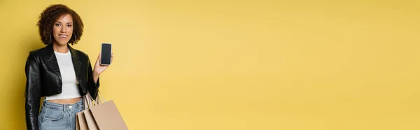 Mulher americana africana feliz em jaqueta de couro segurando sacos de compras e telefone celular com tela em branco no amarelo, banner — Fotografia de Stock