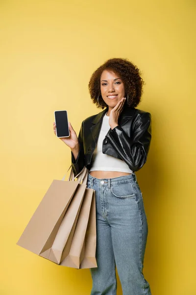 Sorrindo mulher afro-americana em jaqueta de couro segurando sacos de compras e telefone celular com tela em branco no amarelo — Fotografia de Stock