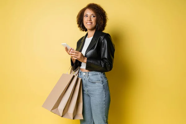 Задоволена афроамериканська жінка в стильній шкіряній куртці, що тримає сумки для покупок та мобільний телефон на жовтому — стокове фото