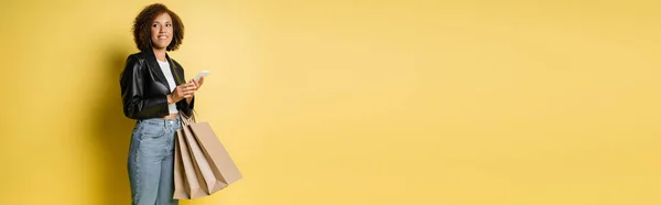Piacere donna afroamericana in elegante giacca di pelle con borse della spesa e telefono cellulare su giallo, banner — Foto stock