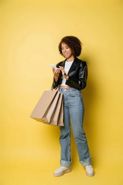 Longitud completa de la mujer afroamericana feliz en la chaqueta de cuero elegante celebración de bolsas de compras y el uso de teléfono móvil en amarillo - foto de stock