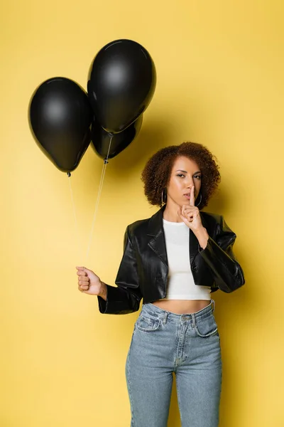 African american woman in leather jacket and jeans standing with black balloons and showing hush on yellow — Stock Photo