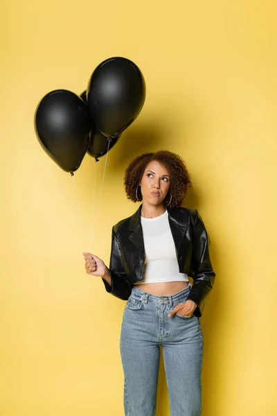 Afrikanisch-amerikanische Frau in Lederjacke und Jeans stehend mit Hand in der Tasche und schwarzen Luftballons auf gelb — Stockfoto