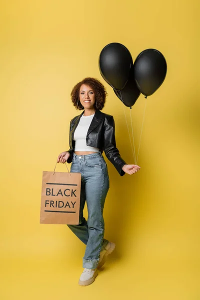 Piena lunghezza di donna afro-americana sorridente in giacca di pelle con in mano venerdì nero shopping bag e palloncini su giallo — Foto stock
