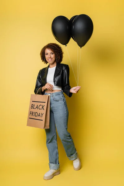 Full length of cheerful african american woman black friday shopping bag and dark balloons on yellow — Stock Photo