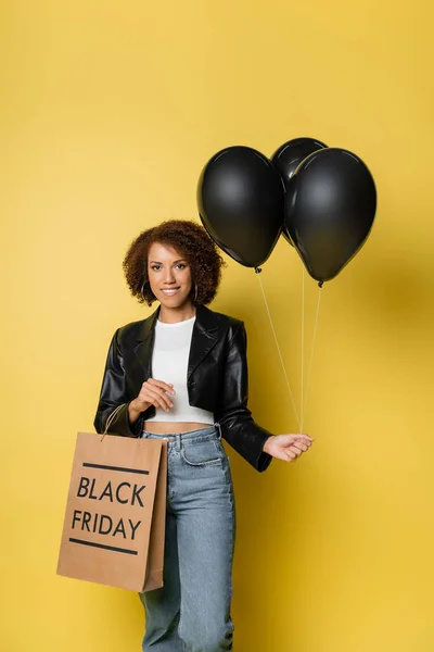 Smiling african american woman in leather jacket holding black friday shopping bag and dark balloons on yellow — Stock Photo