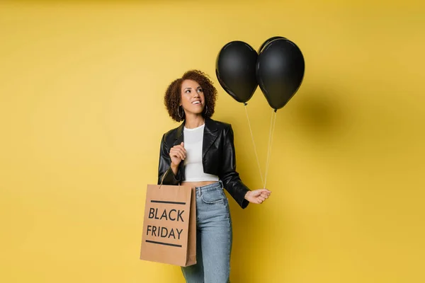 Pleased african american woman in leather jacket holding black friday shopping bag and dark balloons on yellow — Stock Photo