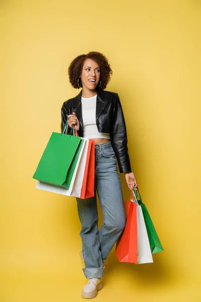 Volle Länge der fröhlichen afrikanisch-amerikanische Frau in Lederjacke hält schwarze Freitag Einkaufstüten auf gelb — Stockfoto