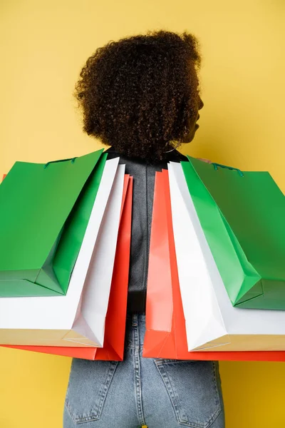 Curly african american woman in denim jeans holding colorful black friday shopping bags on yellow — Stock Photo