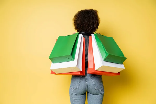 Back view of curly african american woman in stylish blue jeans holding black friday shopping bags on yellow — Stock Photo