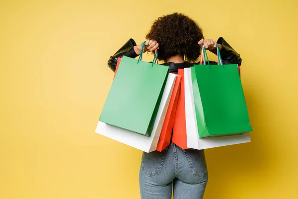 Back view of curly african american woman in blue jeans holding colorful black friday shopping bags on yellow — Stock Photo