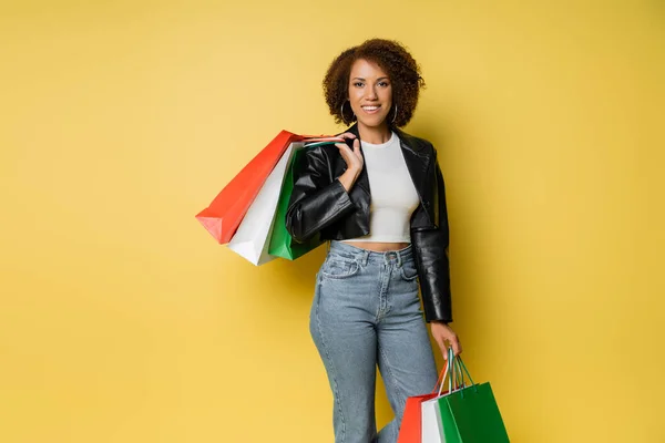 Mulher afro-americana positiva em jeans e jaqueta de couro segurando coloridos sacos de compras preto sexta-feira em amarelo — Fotografia de Stock