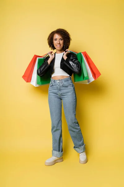 Full length of happy african american woman in jeans and leather jacket holding colorful black friday shopping bags on yellow — Stock Photo