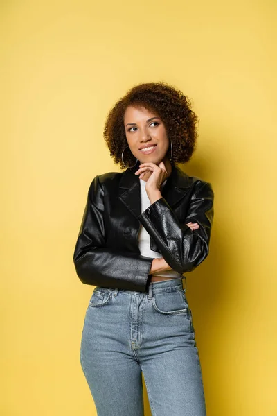 Dreamy african american woman in jeans and leather jacket dreaming and looking away on yellow — Stock Photo