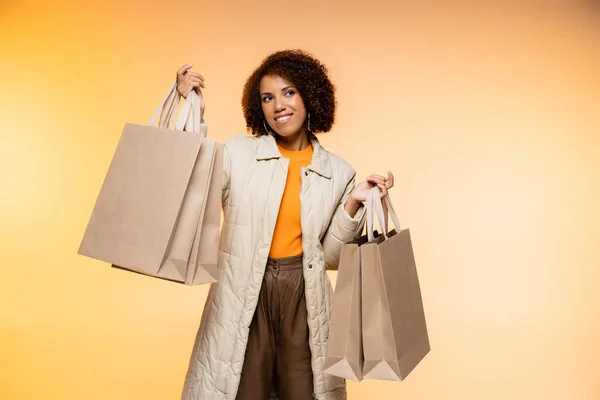 Mujer afroamericana positiva en abrigo sosteniendo bolsas de compras de viernes negro en naranja - foto de stock