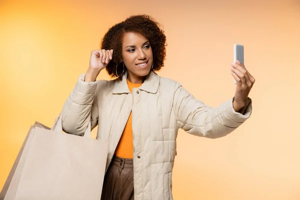 Mujer afroamericana positiva con abrigo sosteniendo bolsas de compras de viernes negro y tomando selfie en naranja - foto de stock