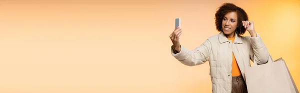 Positive african american woman in coat holding black friday shopping bags and taking selfie on orange, banner — Stock Photo