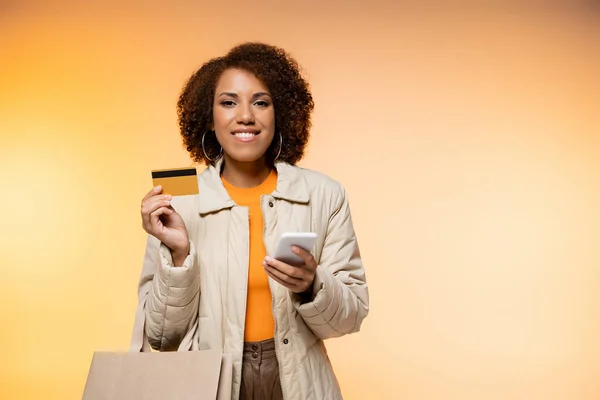 Heureuse femme afro-américaine en manteau tenant smartphone et carte de crédit tout en étant debout avec sac à provisions sur orange — Photo de stock
