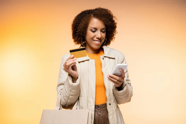 Cheerful african american woman in coat holding smartphone and credit card while standing with shopping bag on orange — Stock Photo