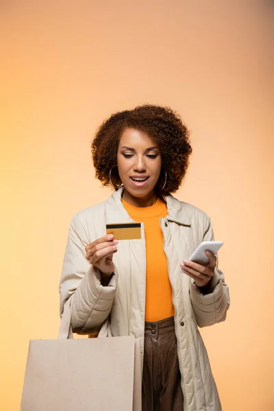 Sorrindo mulher afro-americana em casaco segurando smartphone e cartão de crédito, enquanto de pé com saco de compras em laranja — Fotografia de Stock