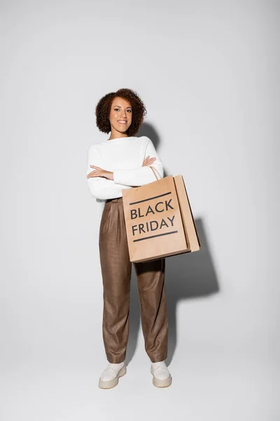 Comprimento total de mulher americana africana feliz em outfit outfit segurando sacos de compras e de pé com braços cruzados em cinza — Fotografia de Stock