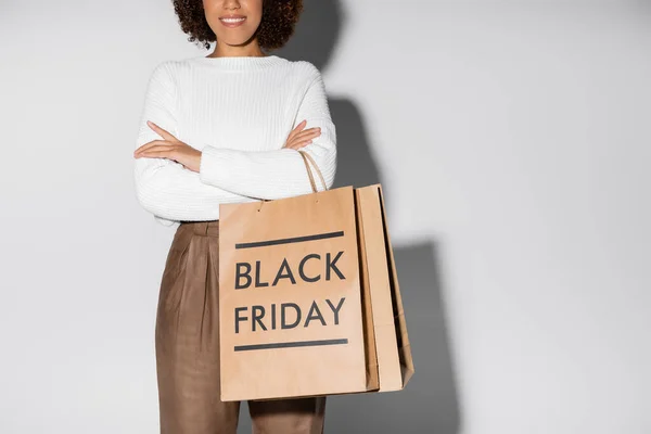 Vue recadrée de la femme afro-américaine heureuse en tenue automnale tenant des sacs à provisions et debout avec les bras croisés sur gris — Photo de stock