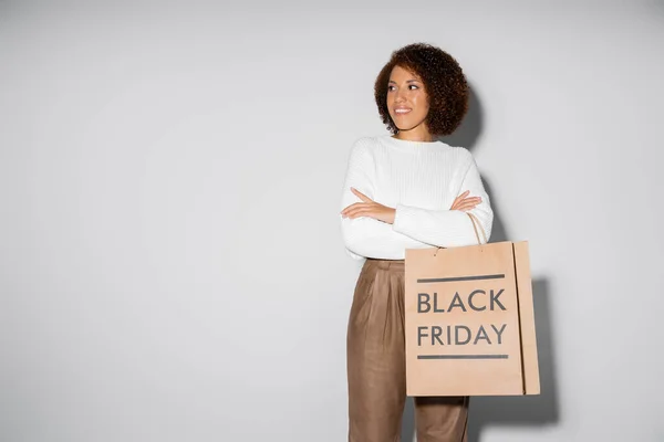 Feliz mulher afro-americana em roupa outnal segurando sacos de compras e de pé com os braços cruzados em cinza — Fotografia de Stock