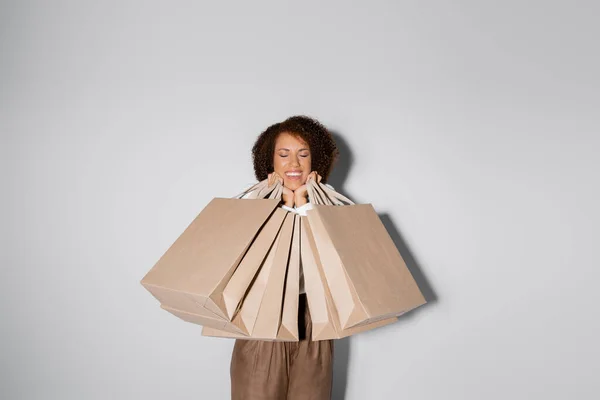 Cheerful african american woman with closed eyes holding shopping bags on grey — Stock Photo