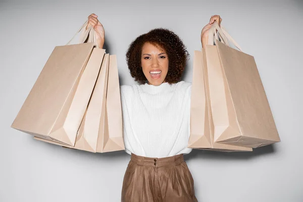 Donna afro-americana stupita in abito autunnale che tiene borse della spesa sul grigio — Foto stock