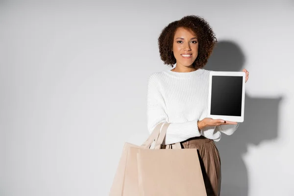 Mulher americana africana feliz em outfit outfit segurando sacos de compras e tablet digital com tela em branco no cinza — Fotografia de Stock