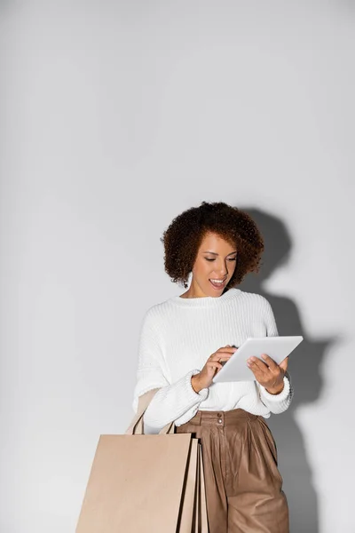 Mujer afroamericana emocionada en traje otoñal sosteniendo bolsas de compras y tableta digital en gris - foto de stock
