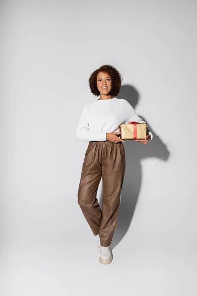 Full length of smiling african american woman in autumnal outfit holding wrapped gift box on grey — Stock Photo