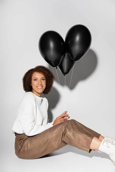 Cheerful african american woman in autumnal outfit holding black balloons on grey — Stock Photo