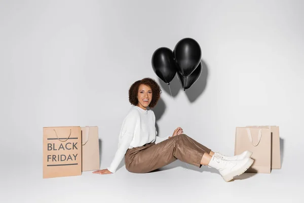 Cheerful african american woman in autumnal outfit holding black balloons near shopping bags on grey — Stock Photo