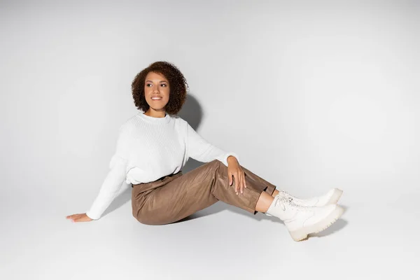 Femme afro-américaine gaie en tenue automnale assis et regardant loin sur gris — Photo de stock