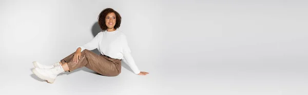 Femme afro-américaine gaie en tenue automnale assis et regardant loin sur gris, bannière — Photo de stock