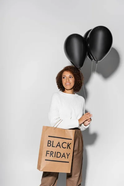 Mulher americana africana alegre em outfit outfit segurando saco de compras e balões pretos em cinza — Fotografia de Stock