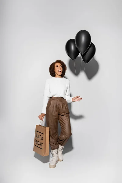 Longitud completa de mujer afroamericana sorprendida en traje otoñal sosteniendo bolsa de compras y globos negros en gris - foto de stock