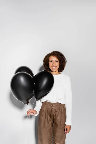 Lächelnde Afroamerikanerin im herbstlichen Outfit mit schwarzen Luftballons auf grau — Stockfoto