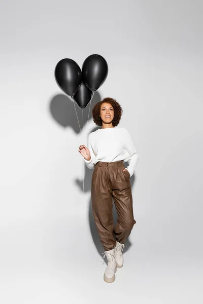 Full length of smiling african american woman in autumnal outfit holding black balloons and posing with hand in pocket on grey — Stock Photo