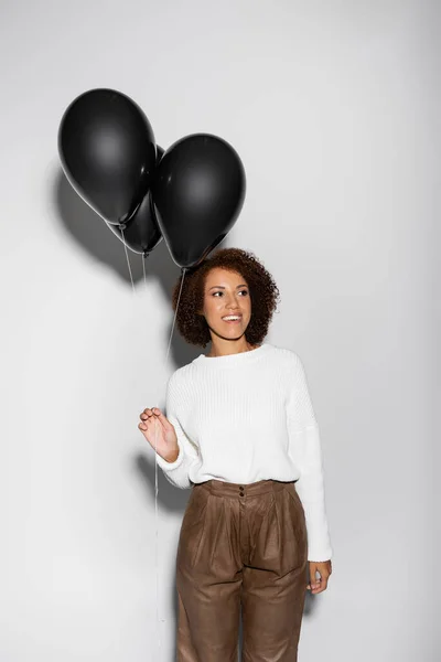 Pleased african american woman in autumnal outfit holding black balloons on grey — Stock Photo