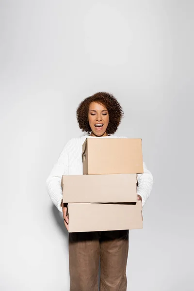 Femme afro-américaine étonnante avec des cheveux bouclés tenant boîtes de livraison sur gris — Photo de stock