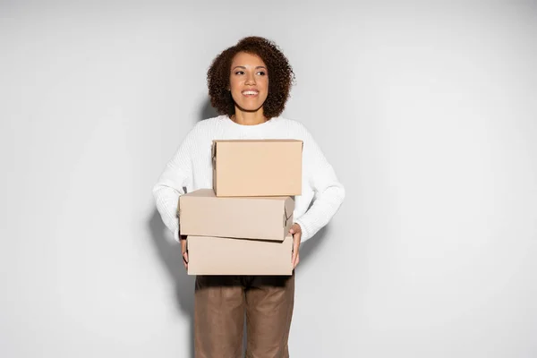 Alegre africana americana mujer con pelo rizado celebración entrega cajas en gris - foto de stock