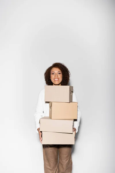 Ravie femme afro-américaine avec des cheveux bouclés tenant boîtes de livraison sur gris — Photo de stock